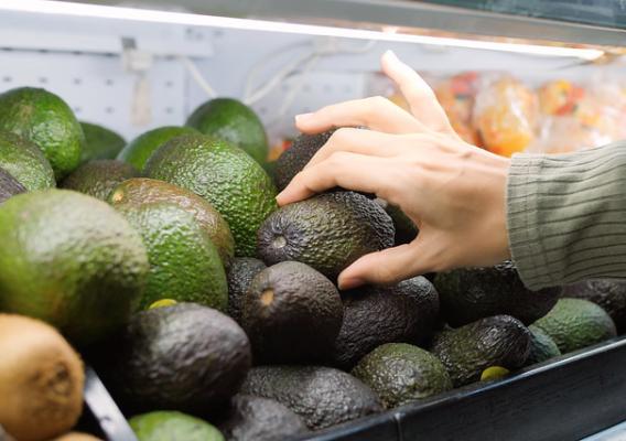 Closeup of a shopper’s hand picking an avocado in a grocery store bin