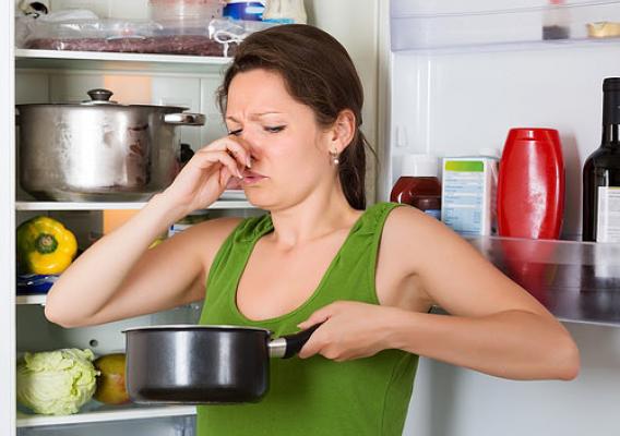 A woman holding her nose at spoiled food in the pot in front of the refrigerator